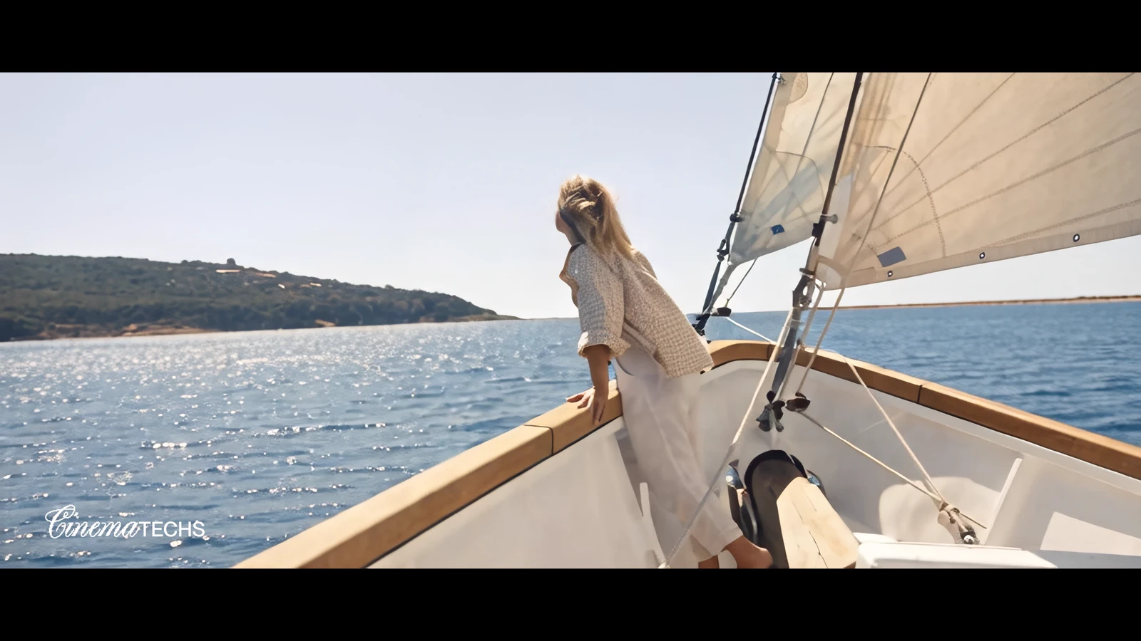 A woman at the bow of a sailboat cutting flat, sunlit water, rigging holding rigid.